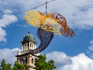 Colorful metal sculpture hangs suspended in air in front of historic building